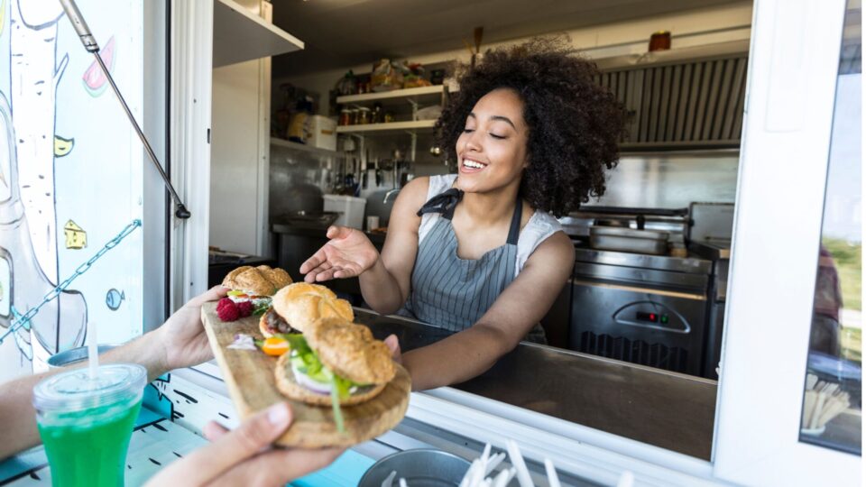 A woman inside a food truck serves an unseen custom a plate of food featuring three small sandwiches