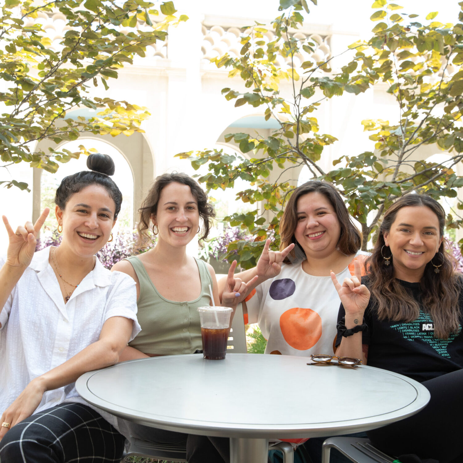 four women sitting at table, smiling and holding up hook 'em hand signs