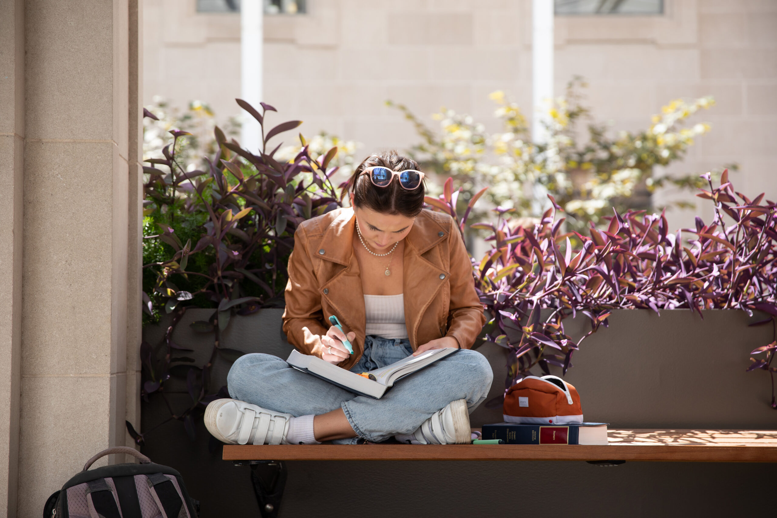 A woman in a brown jacket, blue jeans, and white sneakers sits cross-legged in the courtyard, studying a law book