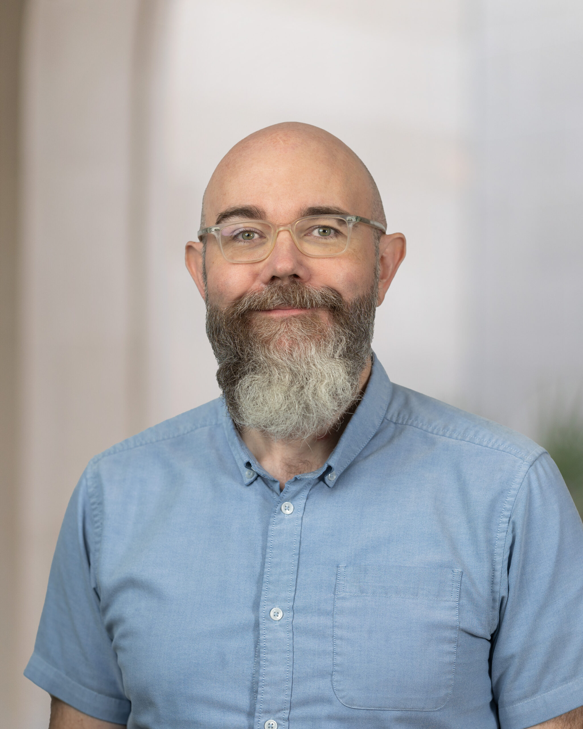 Adam Norwood in a blue Oxford shirt standing in front of a neutral architectural background