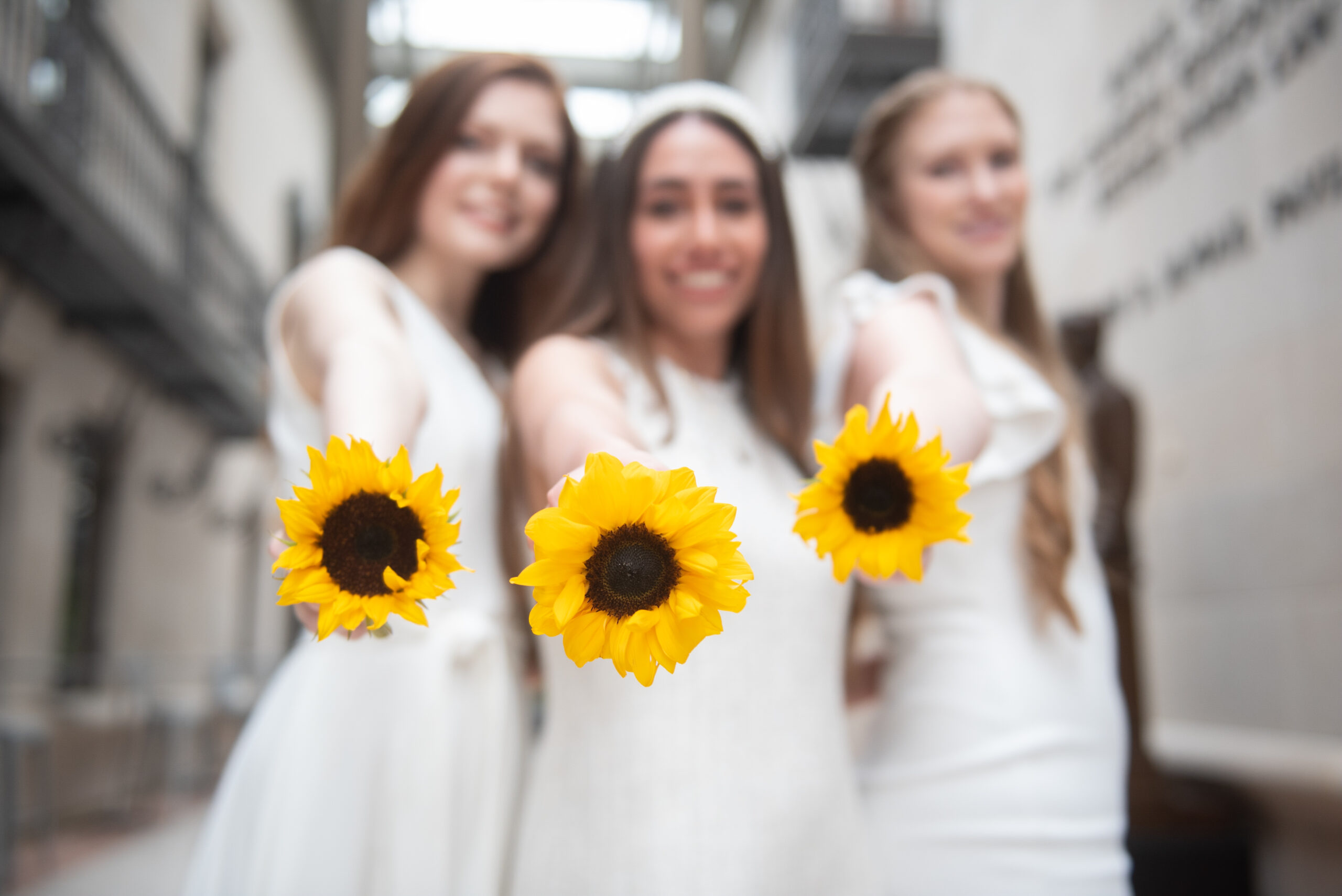 Three graduates in white dresses holding sunflowers.