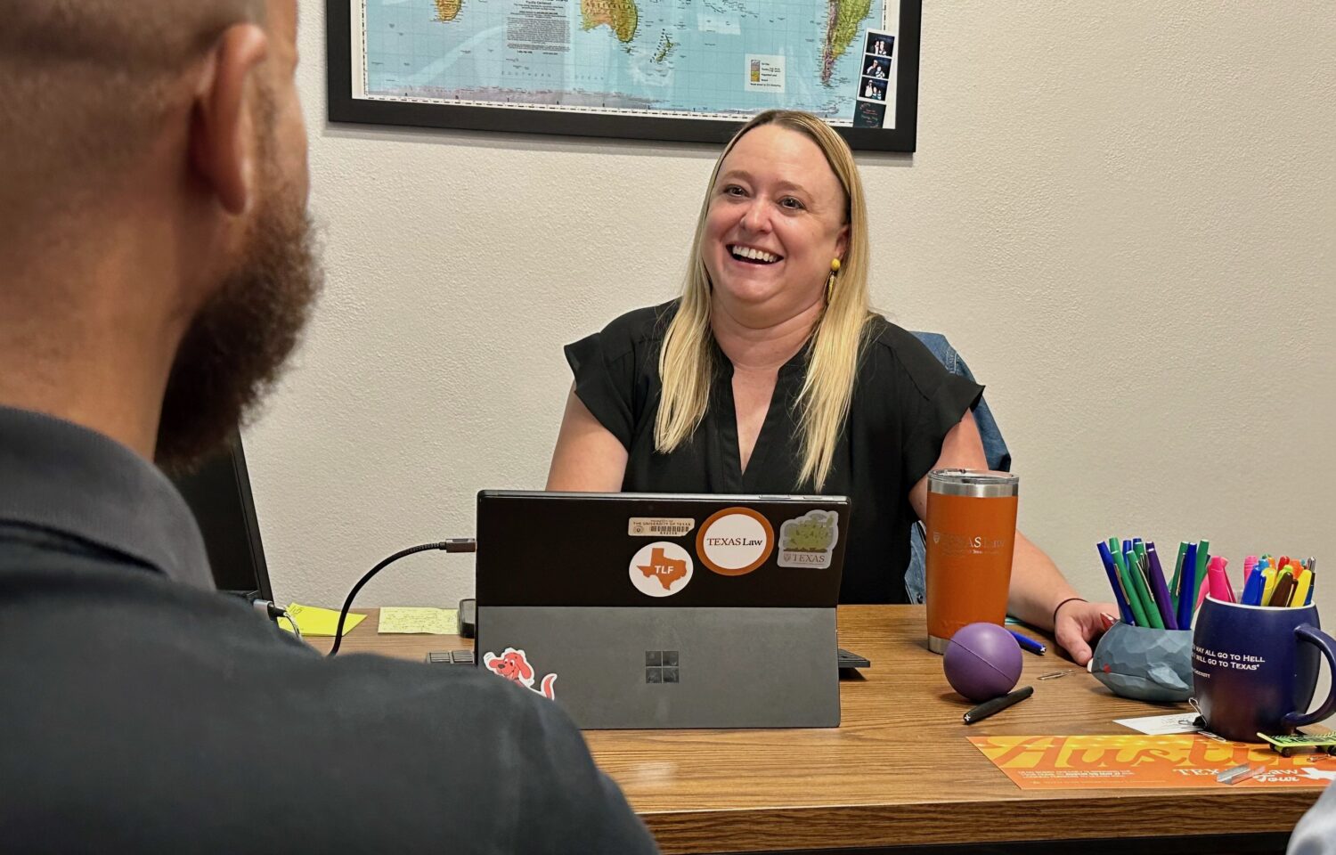 A woman sits at a desk and speaks happily to a student sitting in the foreground with his back to the camera 