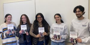 A group of five students stand in the front of a classroom. Each is holding up a book with the words "Sensing Injustice" and "Michael Tigar" on the cover.