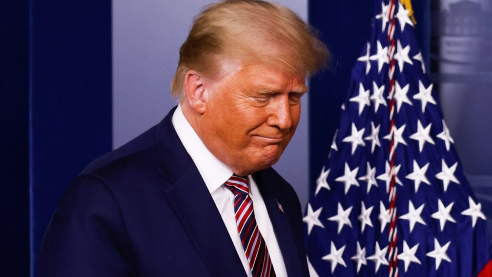 Donald Trump with his eyes closed, looking down, with an American flag behind him.