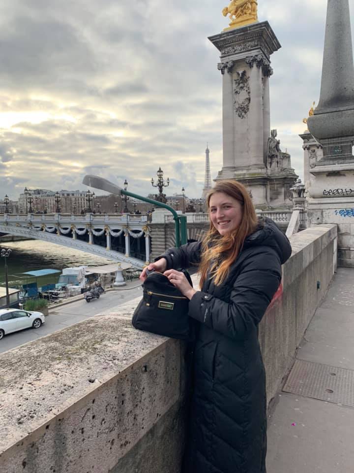 Caroline Burks pictured in a large coat standing in front of stone infrastructures