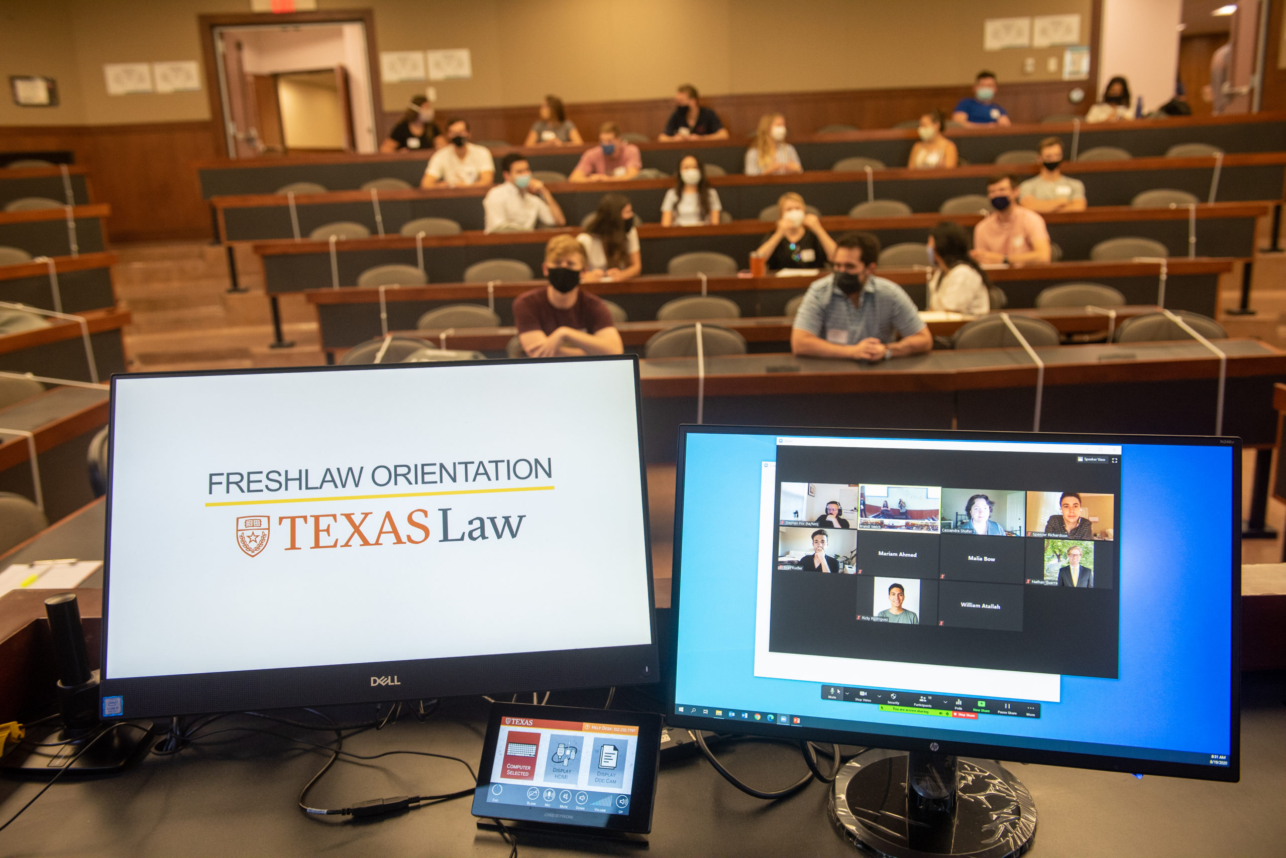 Two computer screens, one showing a Zoom screen, and one showing the Freshlaw Orientation slide, in a room full of students at orientation.