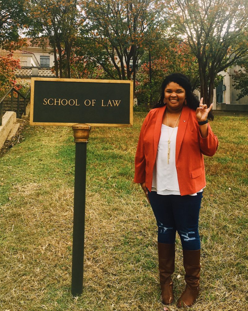 Amber Magee wearing an orange jacket, holding up the 'hook em' emblem, in front of the "School of Law" sign