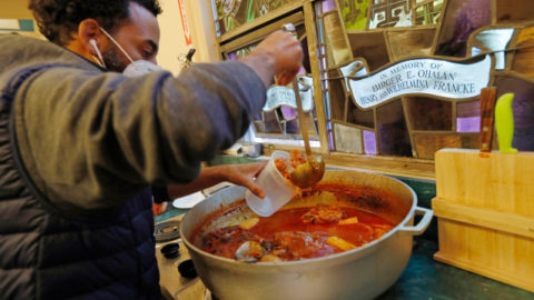 A man wearing a COVID mask ladles stew from a large commercial cooking pot