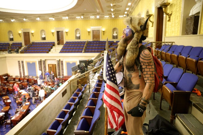 A man holding a United States flag and megaphone, wearing a fur hat with horns and blue face paint is shirtless standing on the top floor of the Senate Chamber during the Capitol Insurrection.