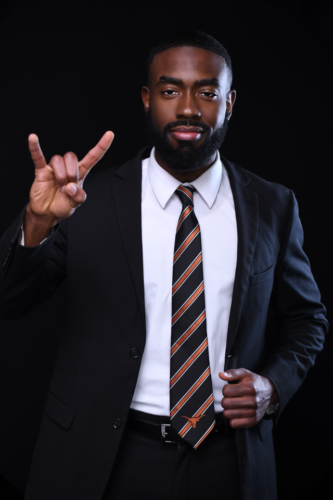 Photograph of Anthony Collier is a dark suit and striped tie and flashing a Hook 'em Horns hand sign