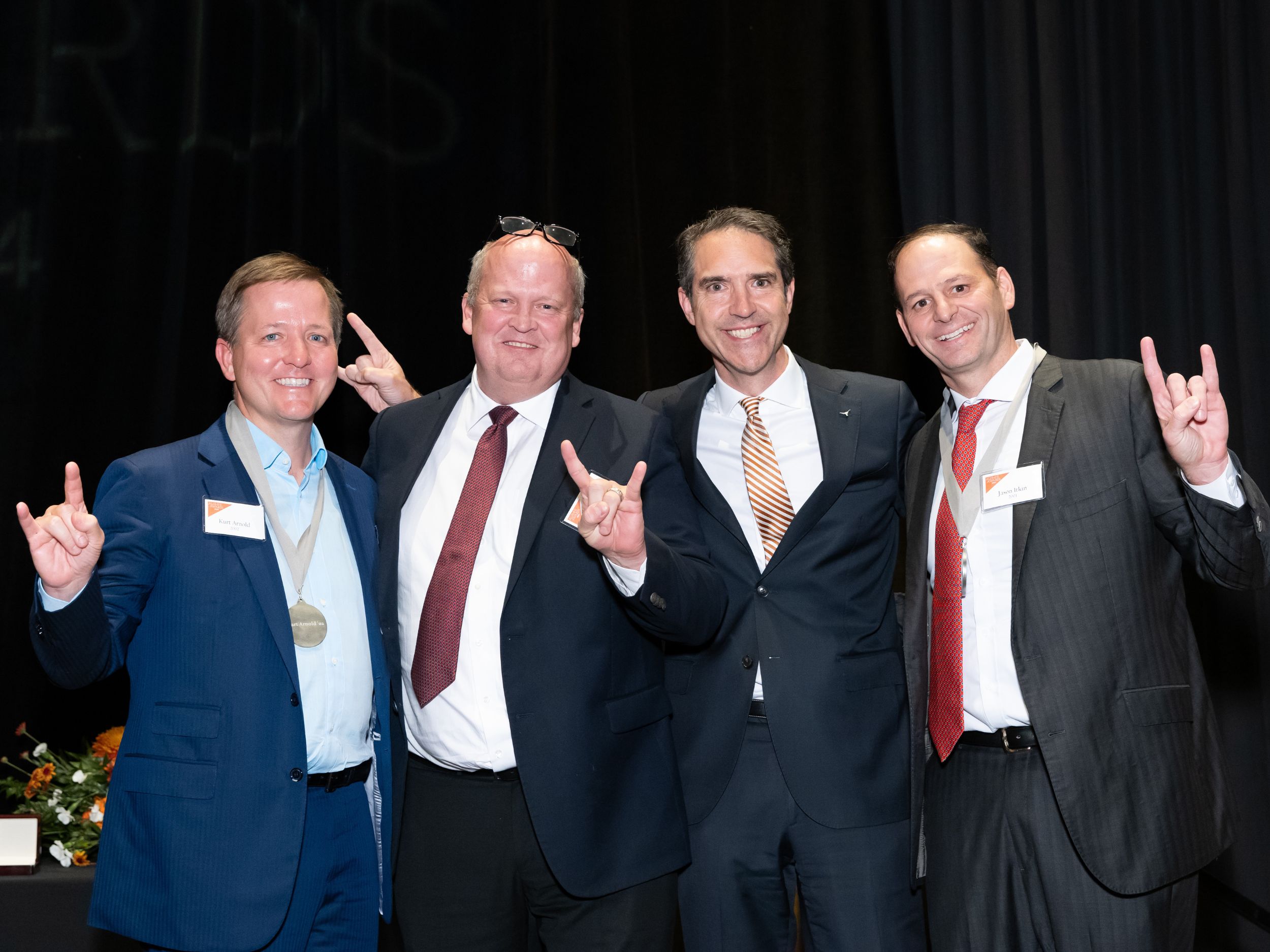 Photo of Honoree Kurt Arnold ’02, award presenter Mikal Watts ’90, Dean Bobby Chesney, and honoree Jason Itkin ’01, wearing suits on stage with black curtain.