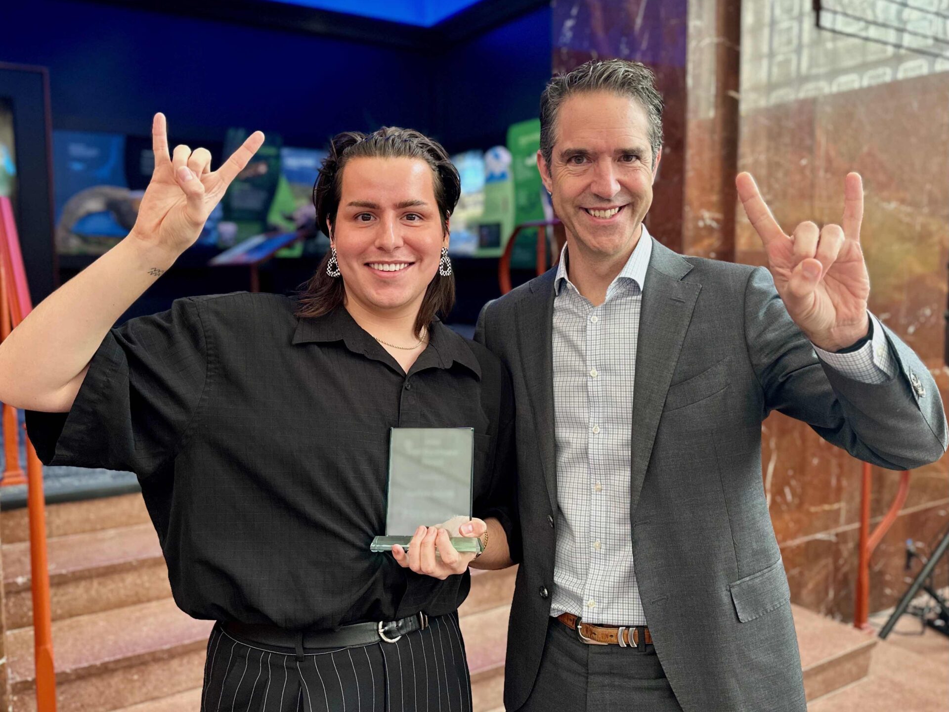 Two people stand side by side, facing the camera, and flashing the "Hook 'em horns" hand sign.