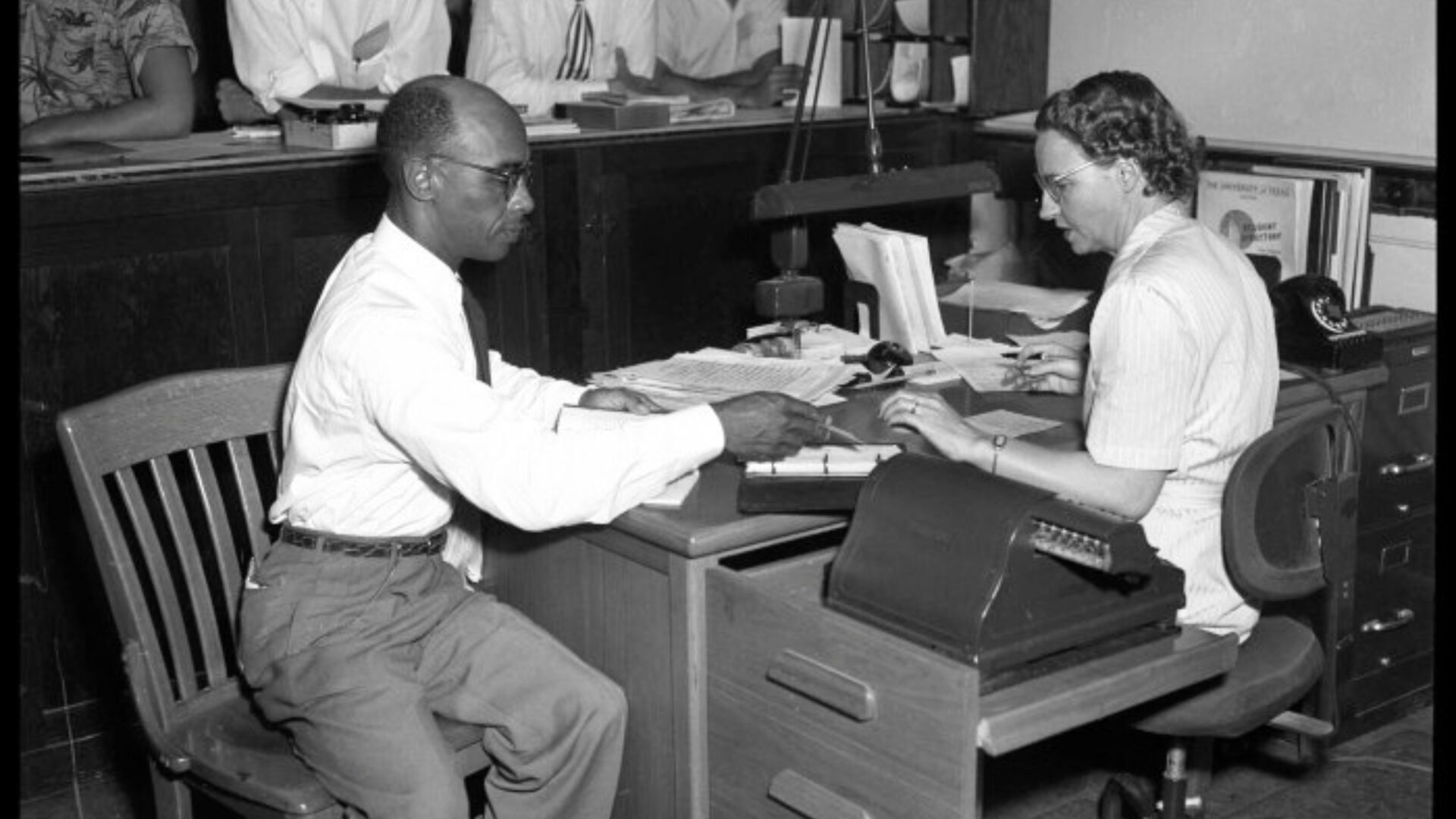 Photograph of Heman Sweatt seated at a table with a woman looking at paperwork, at the Registration Office at the University of Texas at Austin.