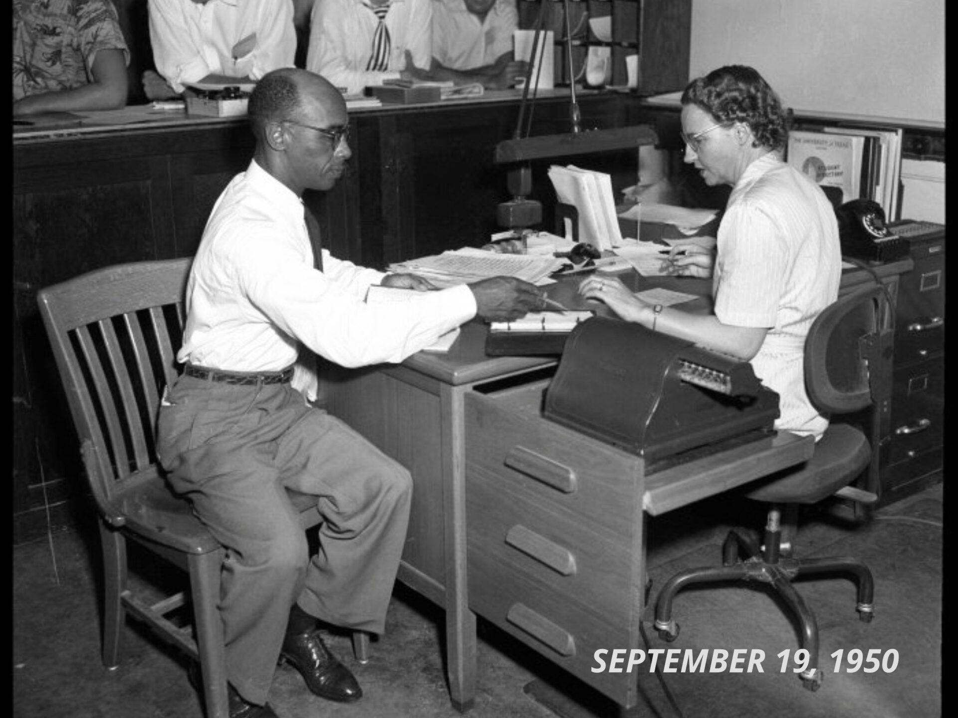 Photograph of Heman Sweatt seated at a table with a woman looking at paperwork, at the Registration Office at the University of Texas at Austin.