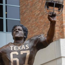 Statue of Julius Whittier from the front, wearing a Texas football jersey with the numbers 67 on front, holding his helmet in the air in his left hand.