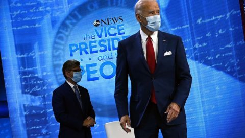 Joe Biden standing in a mask, wearing a red tie and navy suit.