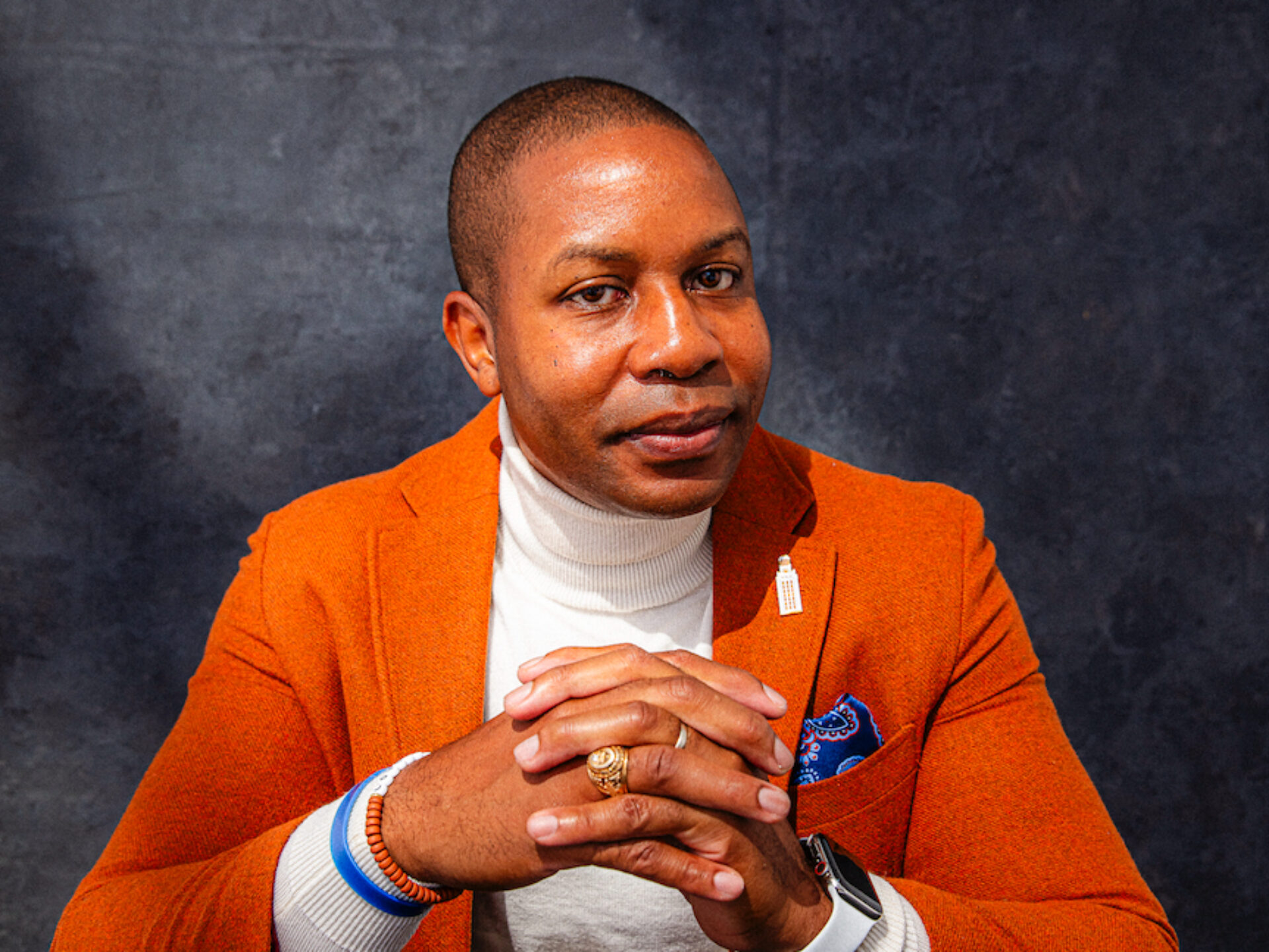 A man seating at a black table, wearing a burnt orange blazer with a white turtleneck underneath and a lapel pin depicting the UT Austin Tower.