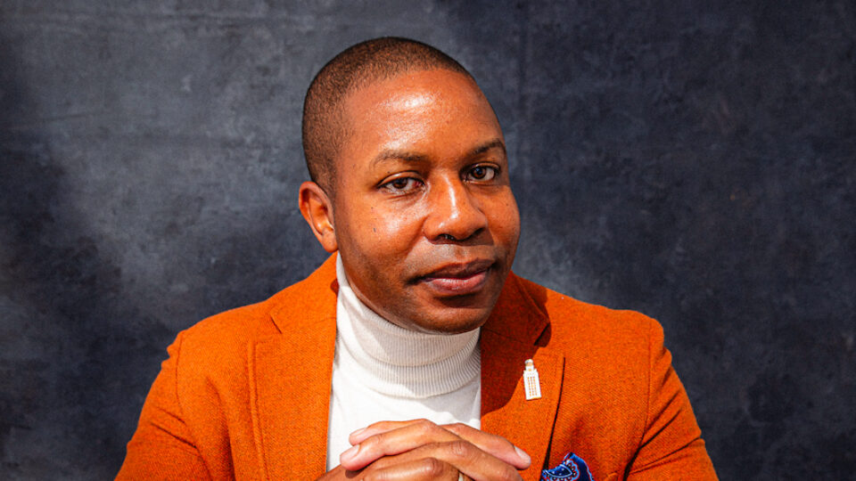 A man seating at a black table, wearing a burnt orange blazer with a white turtleneck underneath and a lapel pin depicting the UT Austin Tower.