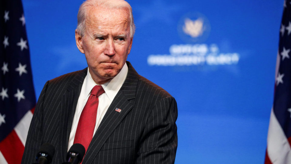 President Joe Biden speaking in front of microphones, wearing a black jacket and a red tie in front of a blue background and American flags.