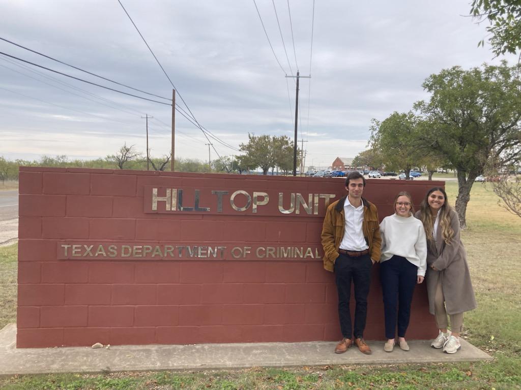 Three law students stand beside the outdoor sign of Hilltop Unit, a Texas correctional facility