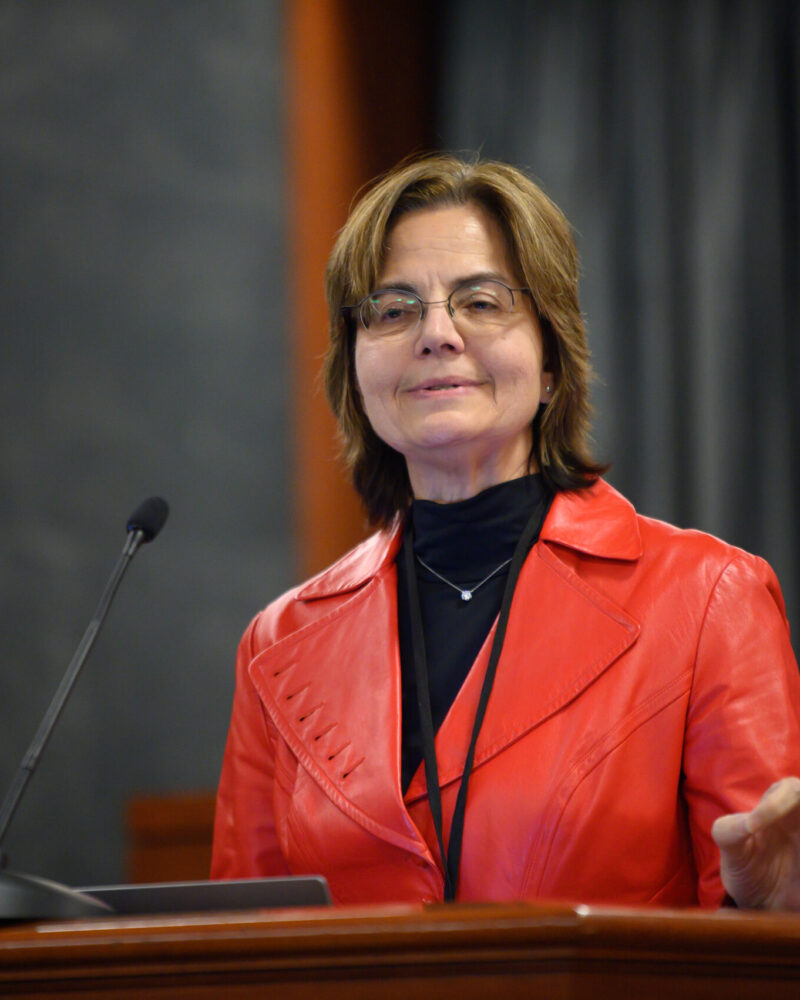 Prof. Lynn Baker stands behind a podium wearing a black shirt underneath a red leather jacket.