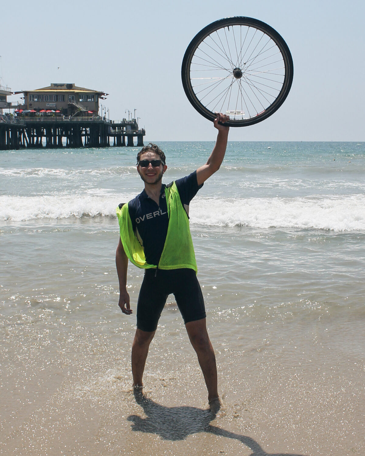 Sam Collins holding bike wheel above his head on a shoreline