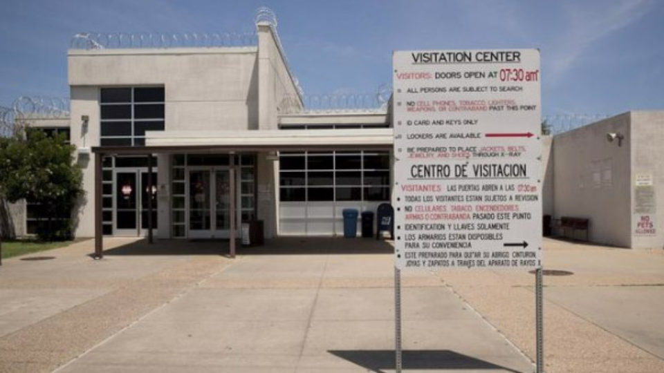 Non-descript building topped with razor wire; a bilingual (English/Spanish) sign in the foreground reads Visitation Center