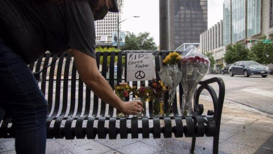 A person leaves an offering among memorial flower bouquets on a street bench, with a sign that reads R.I.P. Garrett Foster