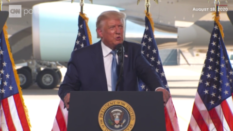 President Trump speaking into a microphone with four American flags behind him.