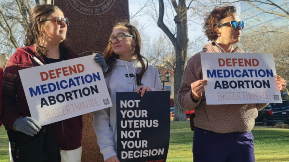 Three women hold up "defend medication abortion" signs.