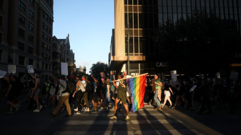 A protest in Texas with individuals crossing the street, holding posters, with one individual in the center holding a large rainbow flag.