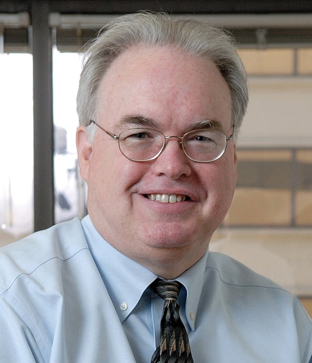 Portrait of Tim Delaney, smiling at the camera wearing glasses and a light blue shirt with a patterned tie.