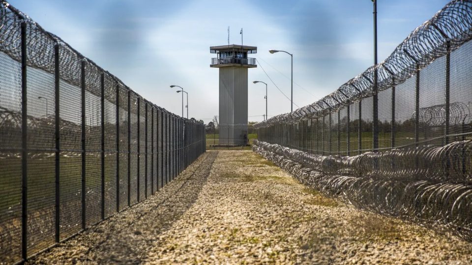 A prison guard tower overlooks a small path surrounded on both sides with fences and razor wire