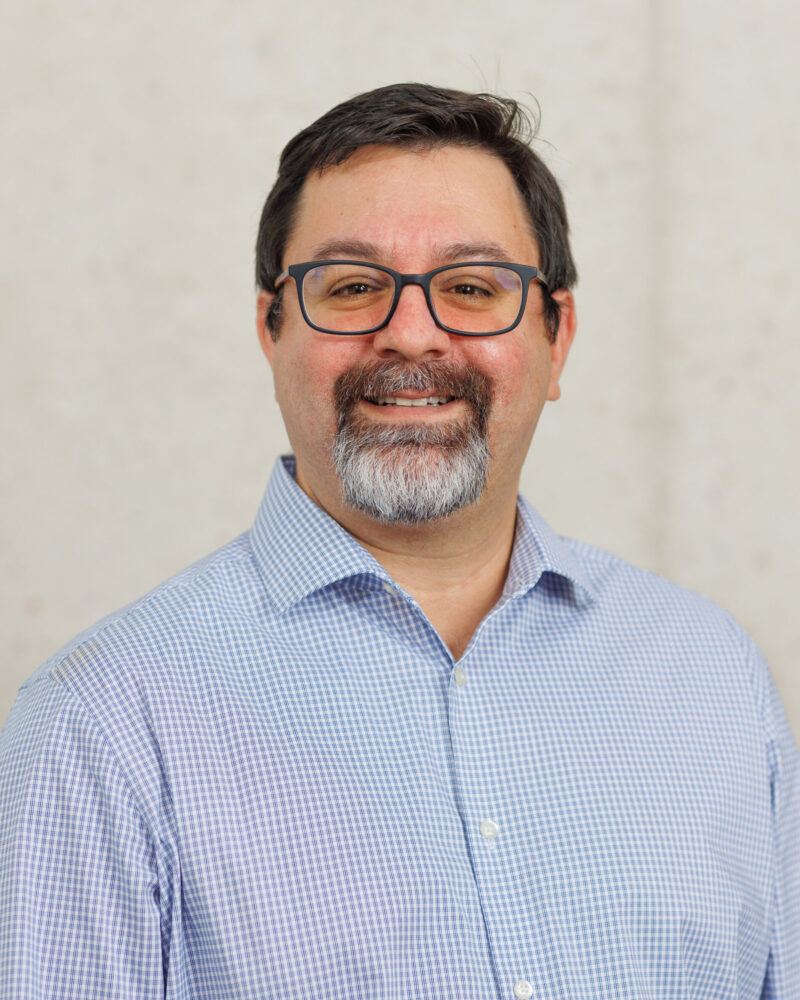 Portrait of Prof. Erik Encarnacion wearing a blue and white checkered button up shirt and glasses.