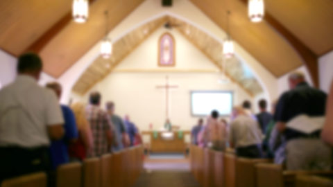 A blurred photo of the inside of a church sanctuary that is filled with people in the pews, and the pastor stands under a large cross at the altar.