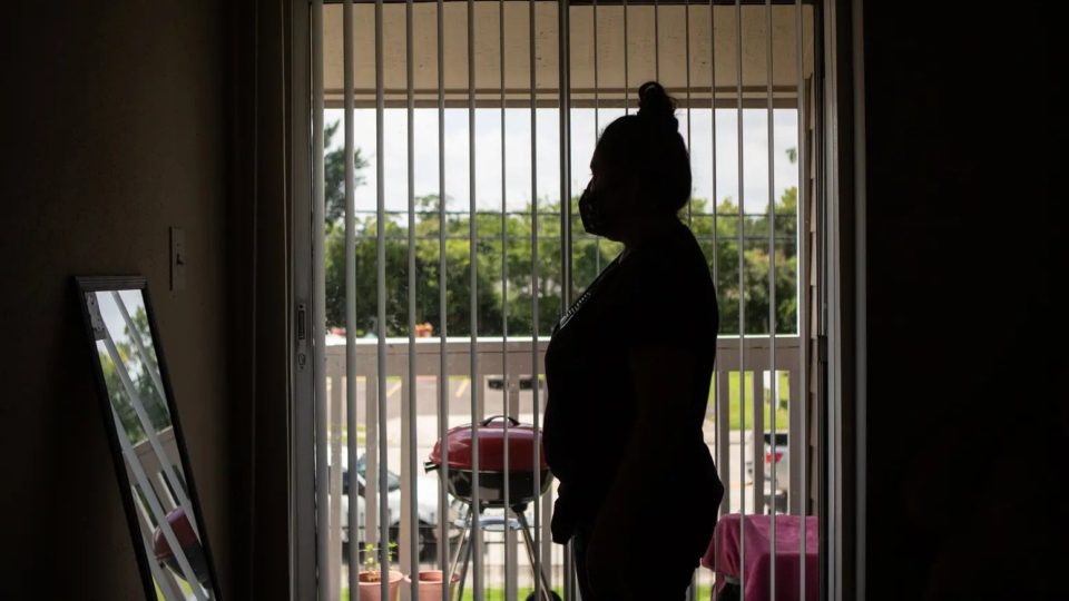 A woman wearing a COVID mask stands in a darkened apartment, in front of sliding glass doors looking onto a patio