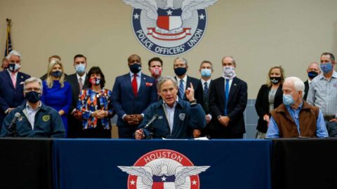 Gov. Greg Abbott, sitting in the center of a large table, talks to reporters during a press conference inside the Houston Police Officer's Union Headquarters, where he signed a "back the blue" pledge. Surrounding him are individuals wearing masks.