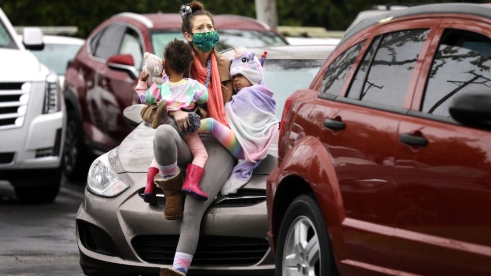 A woman with a concerned look, wearing a COVID-19 mask, sits on the hood of her car in a line of cars, hugging two small children in her lap
