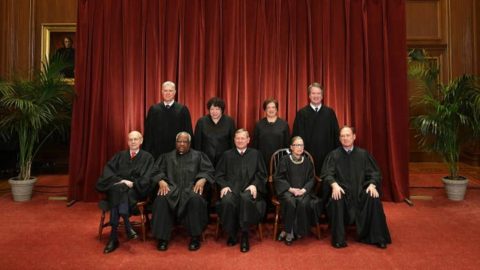 The 9 seated members of the Supreme Court posing in front of a deep red background.
