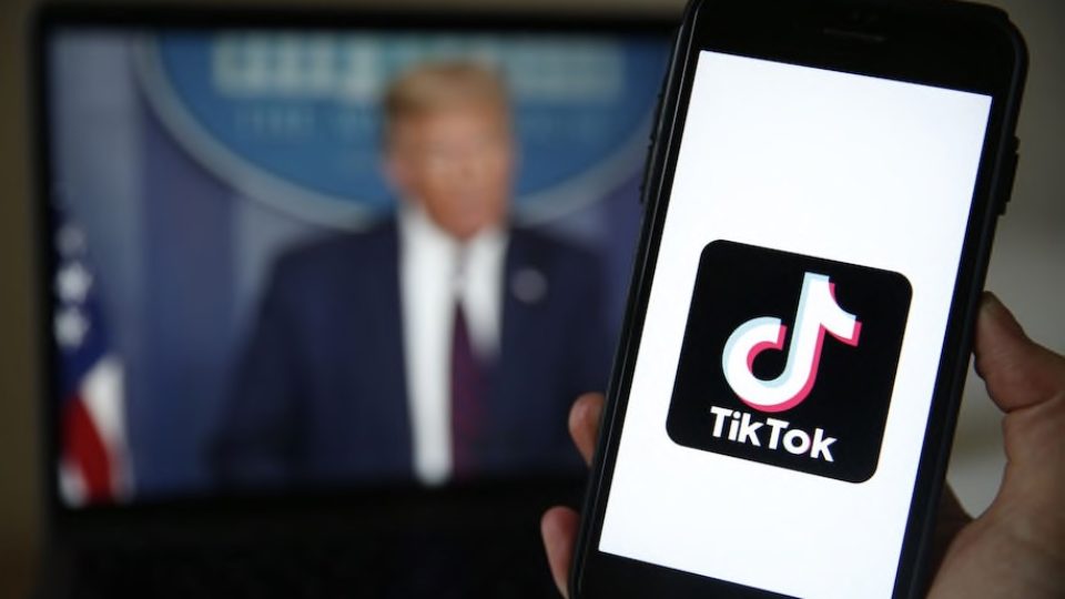 The TikTok app's splash screen held up on a phone in front of a TV image of Donald Trump