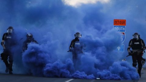 Men wearing riot gear with STATE PATROL insignia walk through a purple cloud of tear gas
