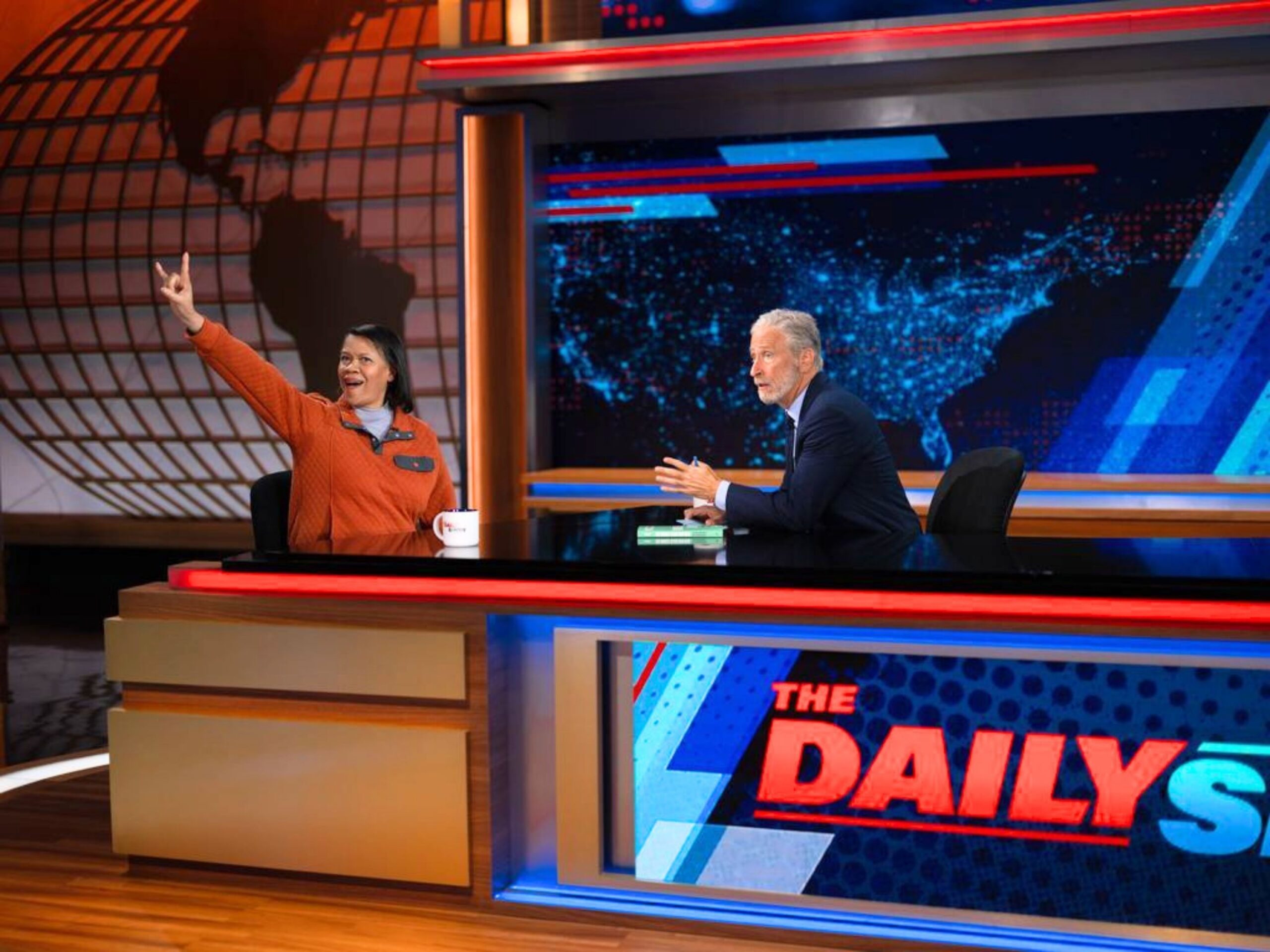 Mechele Dickerson sits next to Jon Stewart on "The Daily Show" showing a hook 'em hand sign.