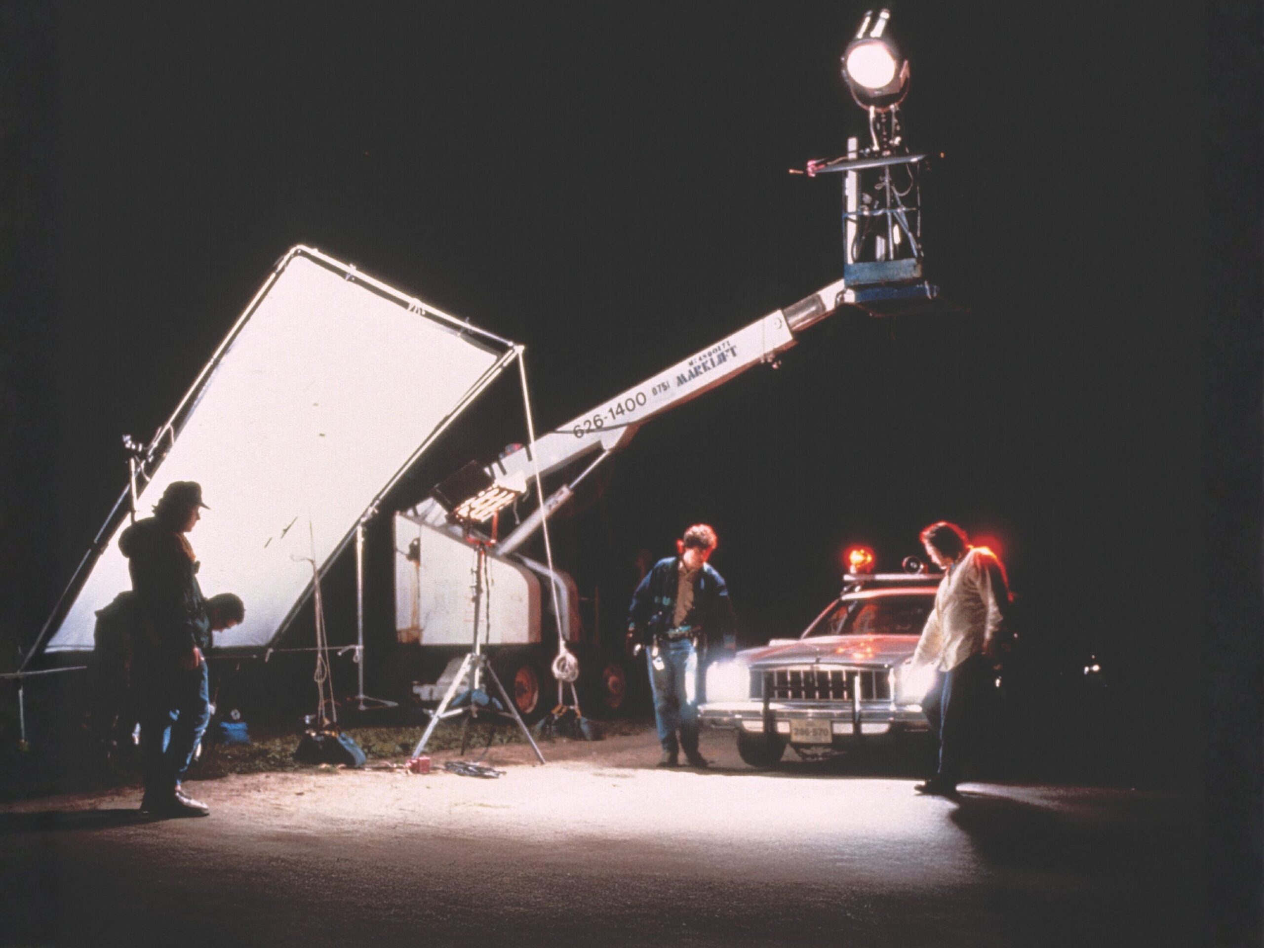 Nighttime film set with a crew working around a car with flashing lights, illuminated by large studio lights and reflectors, while a boom lift holds a bright overhead spotlight.
