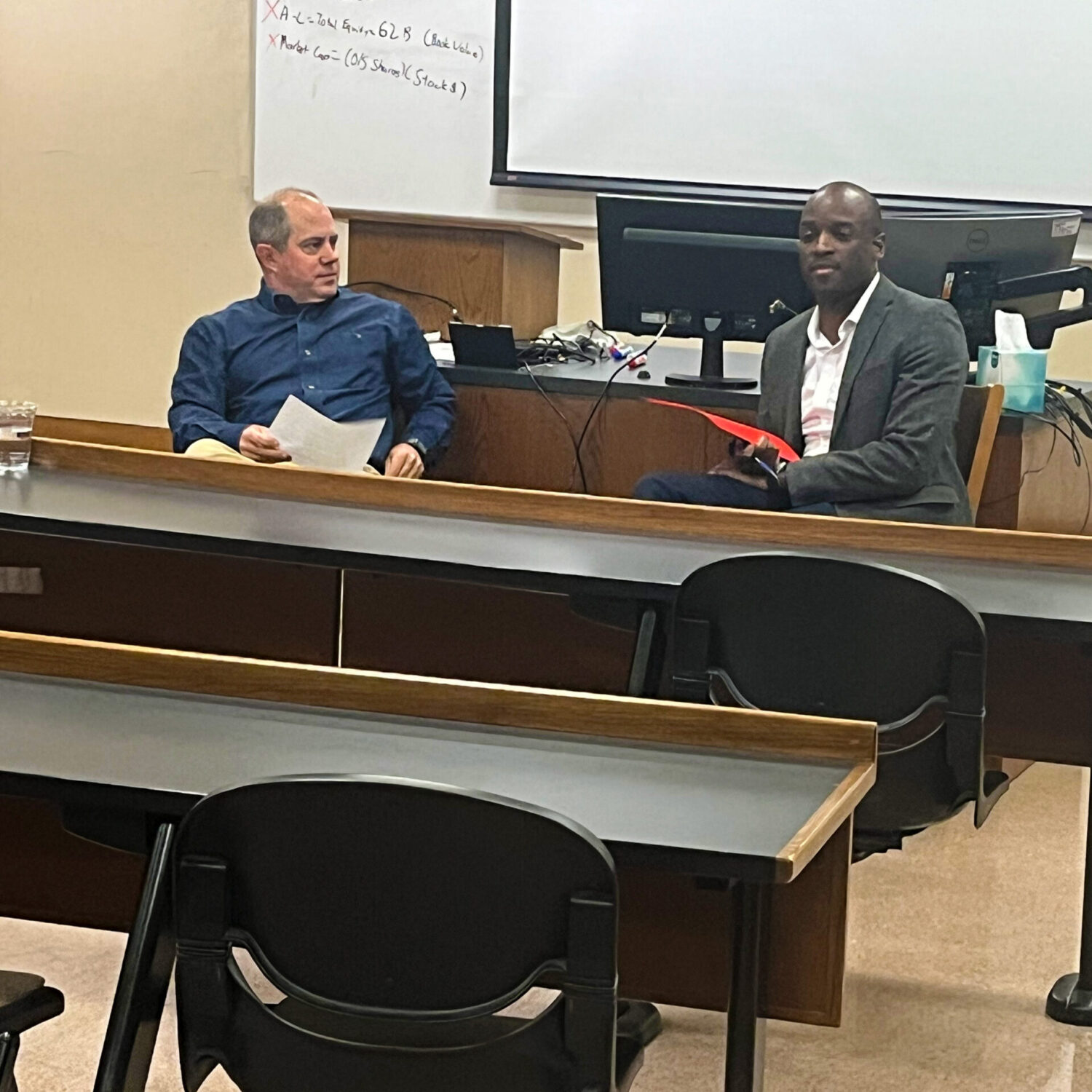 Two men sitting in front of a classroom, facing audience.