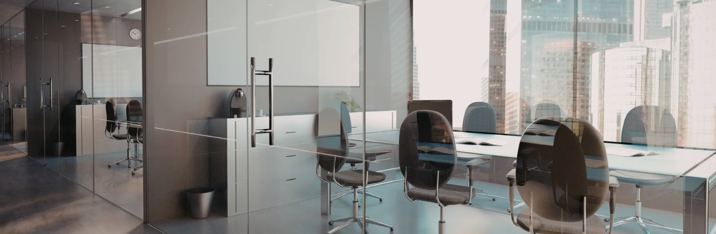 An empty modern conference room with glass walls, a long table surrounded by gray office chairs, and floor-to-ceiling windows overlooking a city skyline.